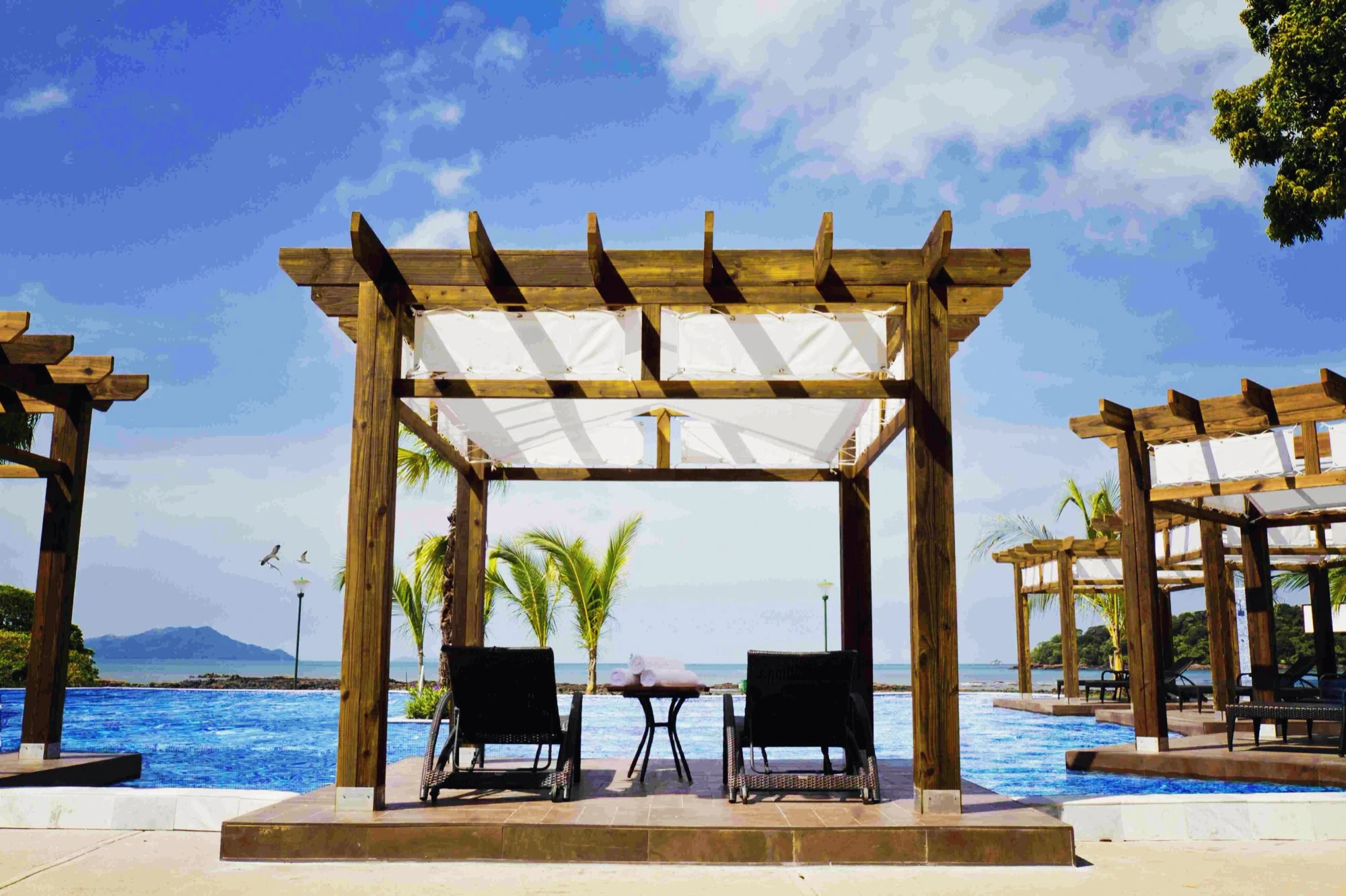 Timber pergolas shade loungers at the pool's edge, the sea and an island beyond